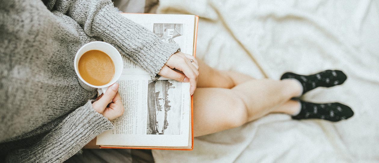 Woman holding a cup of coffee in her right hand reading a book on the bed